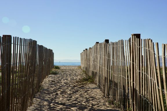 Sandweg zwischen Holzlattenzaun zum Meer bei klarem Himmel.
